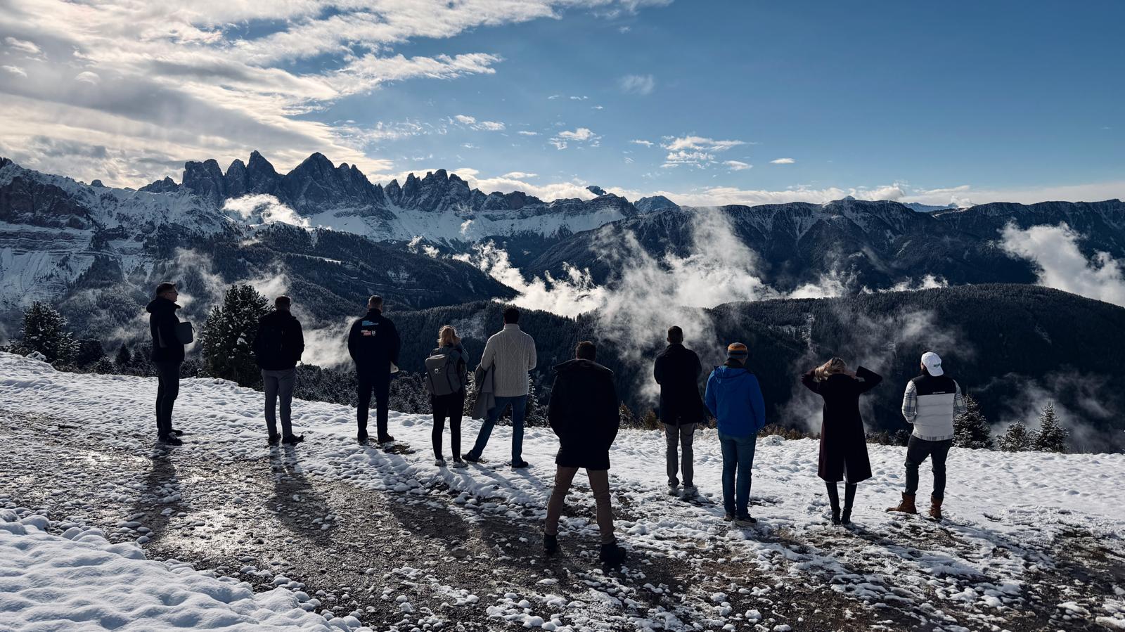 Zehn Teilnehmer stehen von hinten gesehen auf einer verschneiten Anhöhe und blicken auf die Dolomitengipfel über einem Wolkenmeer.