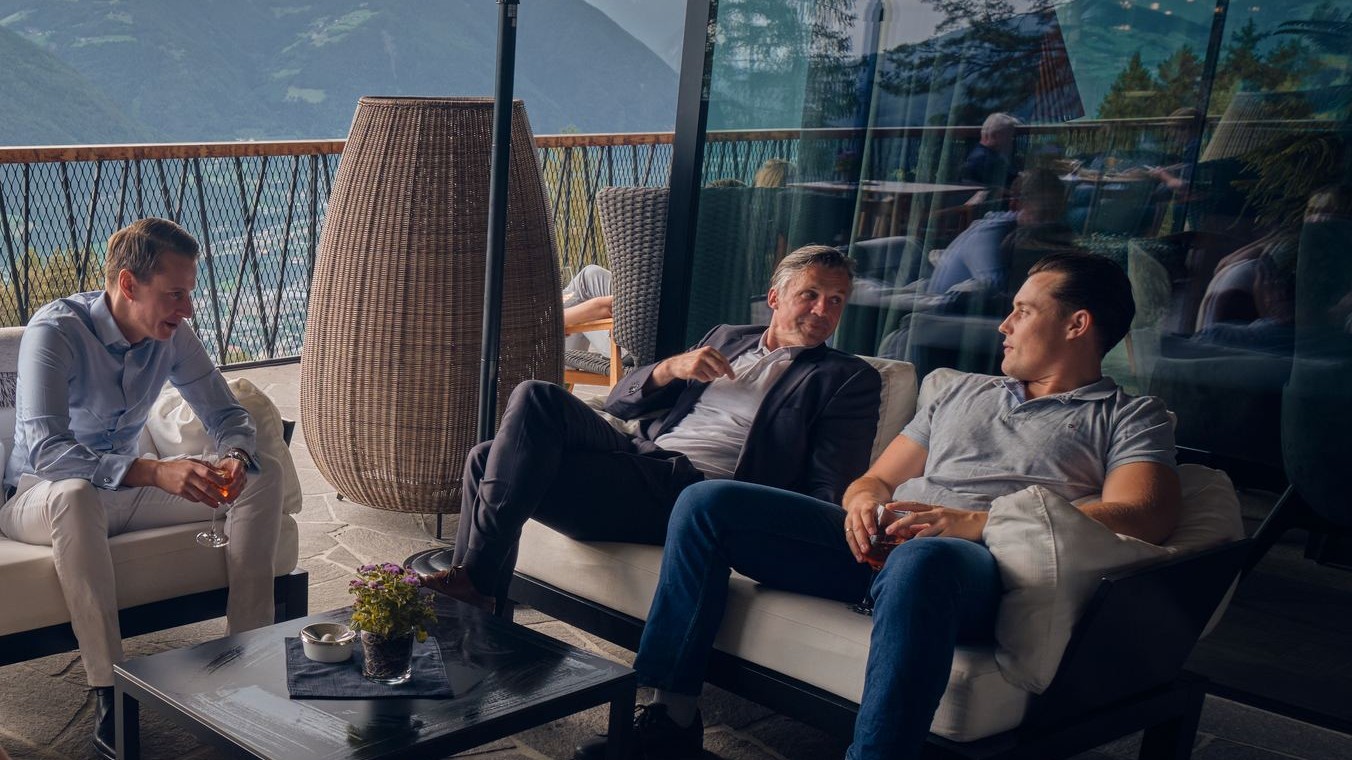 Drei Teilnehmer sitzen auf Sofas auf einer Terrasse mit Bergblick und unterhalten sich.